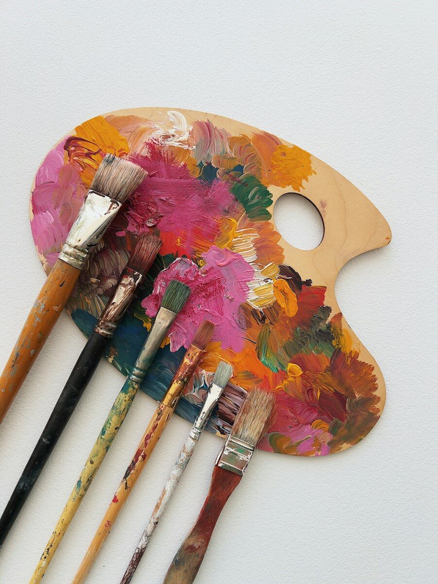 Photo by Jennie Razumnaya Six paintbrushes resting on a wooden palette covered in colorful oil paint